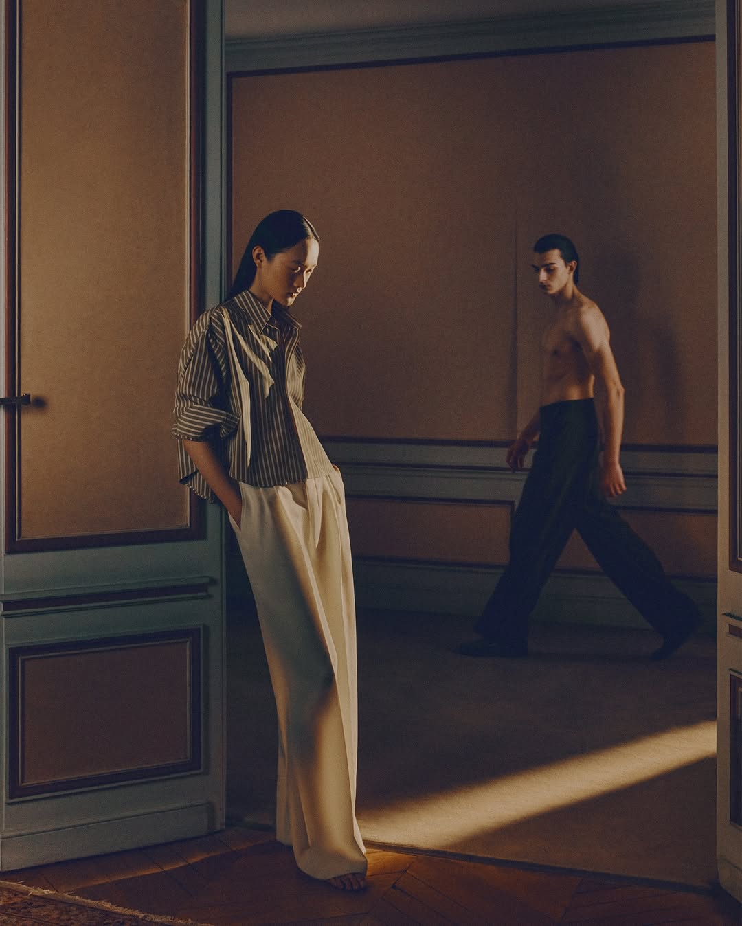 Woman in striped shirt and wide-leg pants stands in doorway as shirtless man in dark pants walks into room with warm lighting.