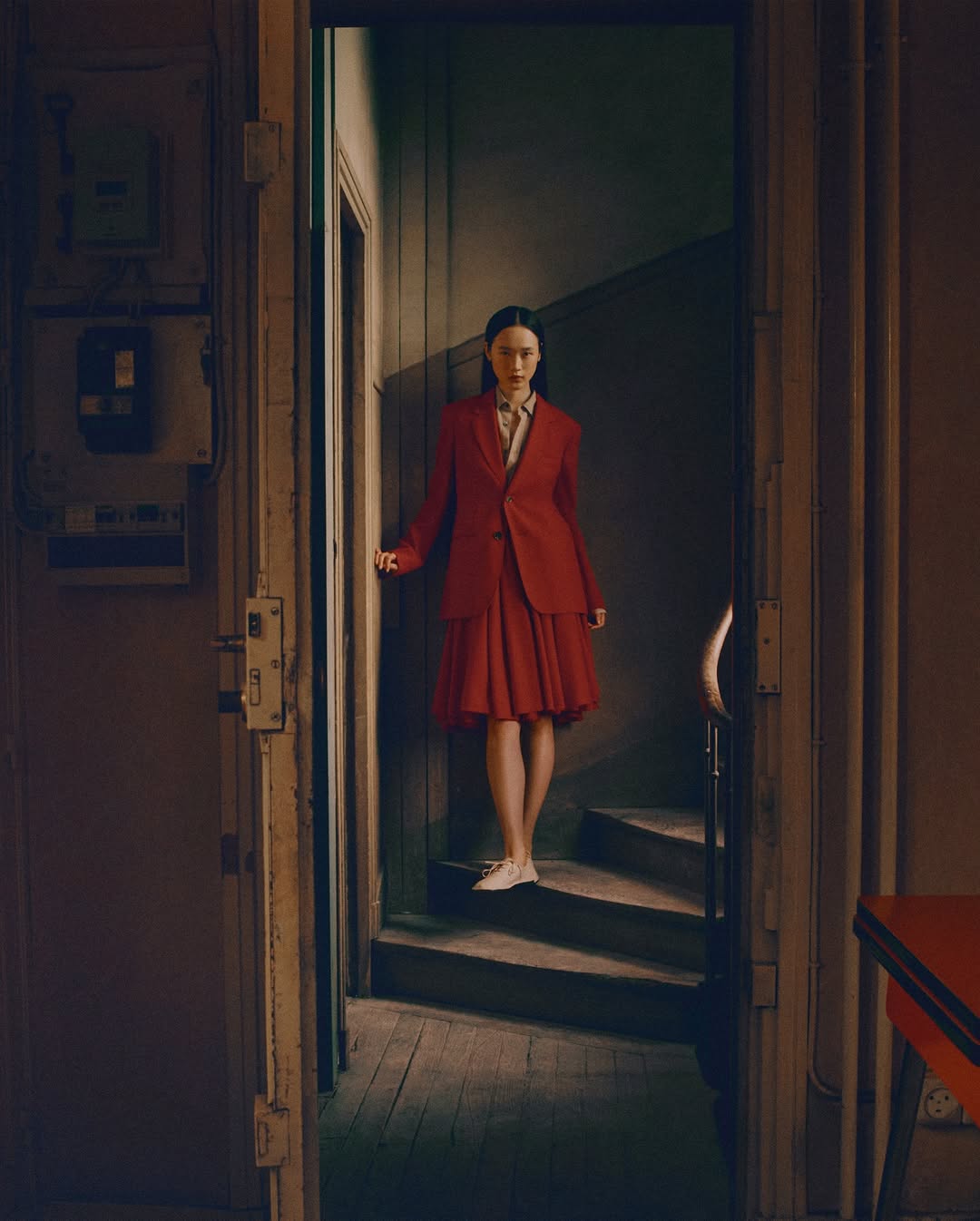 Woman in red suit walks down staircase, viewed through doorway, in vintage interior with muted colors, soft lighting, and aged textures.