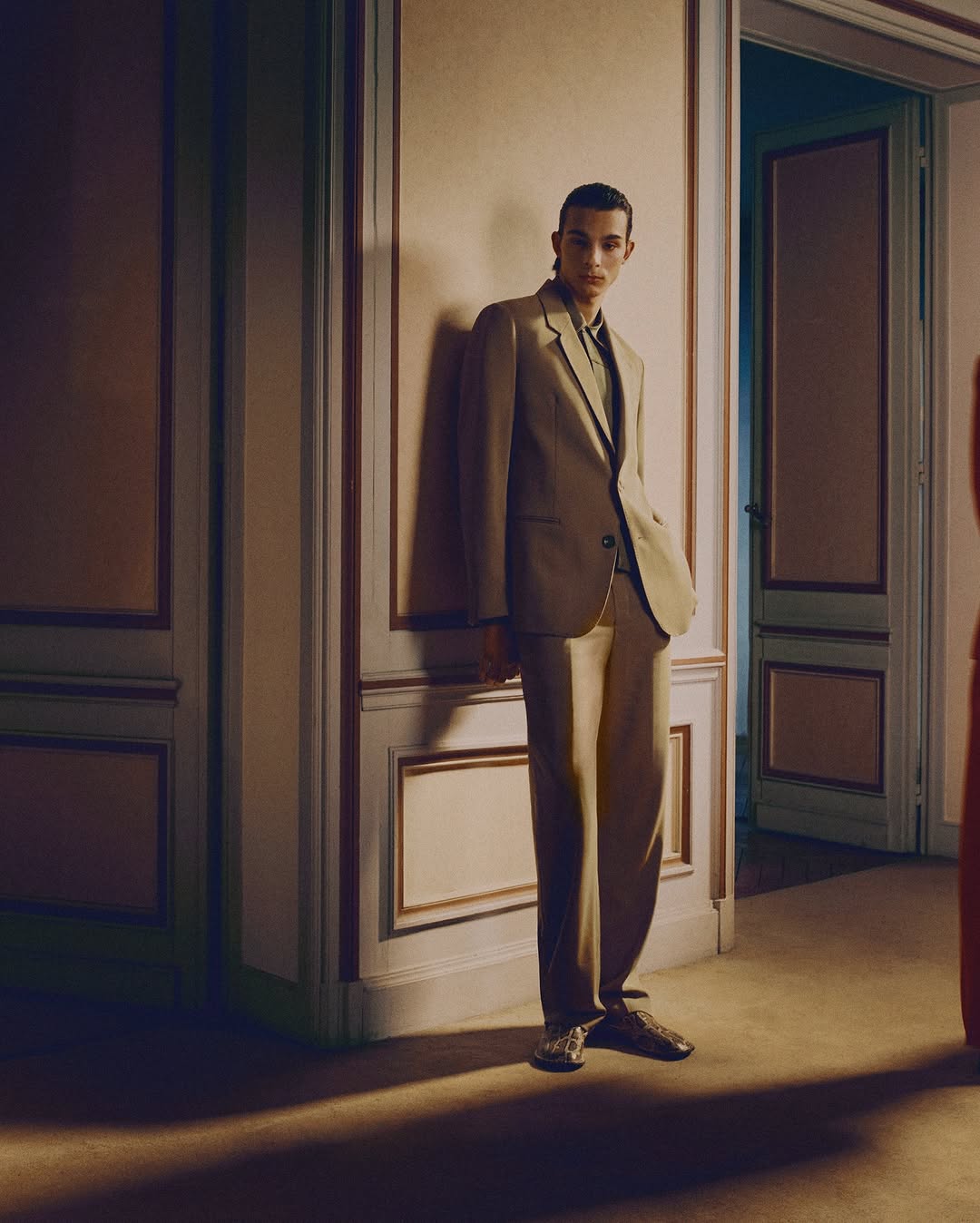 A young adult man in a stylish beige suit poses in an elegant indoor space with a doorway, showcasing a modern fashion aesthetic.