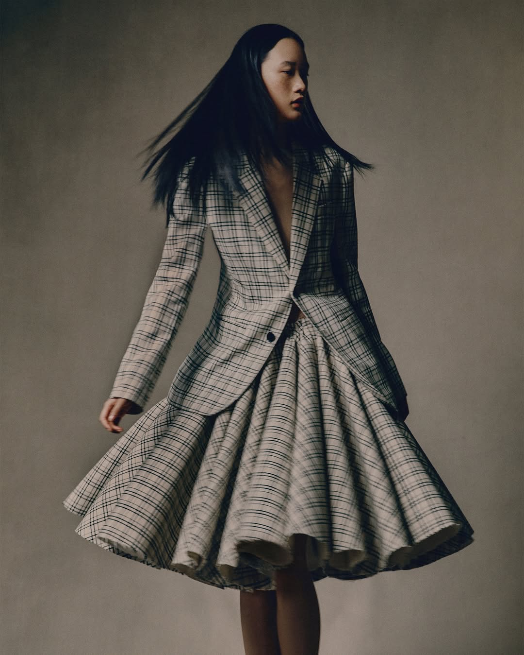 Model with long dark hair wearing a plaid blazer and flared pleated skirt against a neutral studio backdrop.
