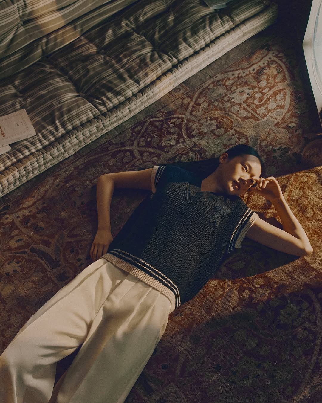 Woman lies on a patterned rug in a sleeveless sweater and pants, gazing off camera, with a striped mattress in the background.