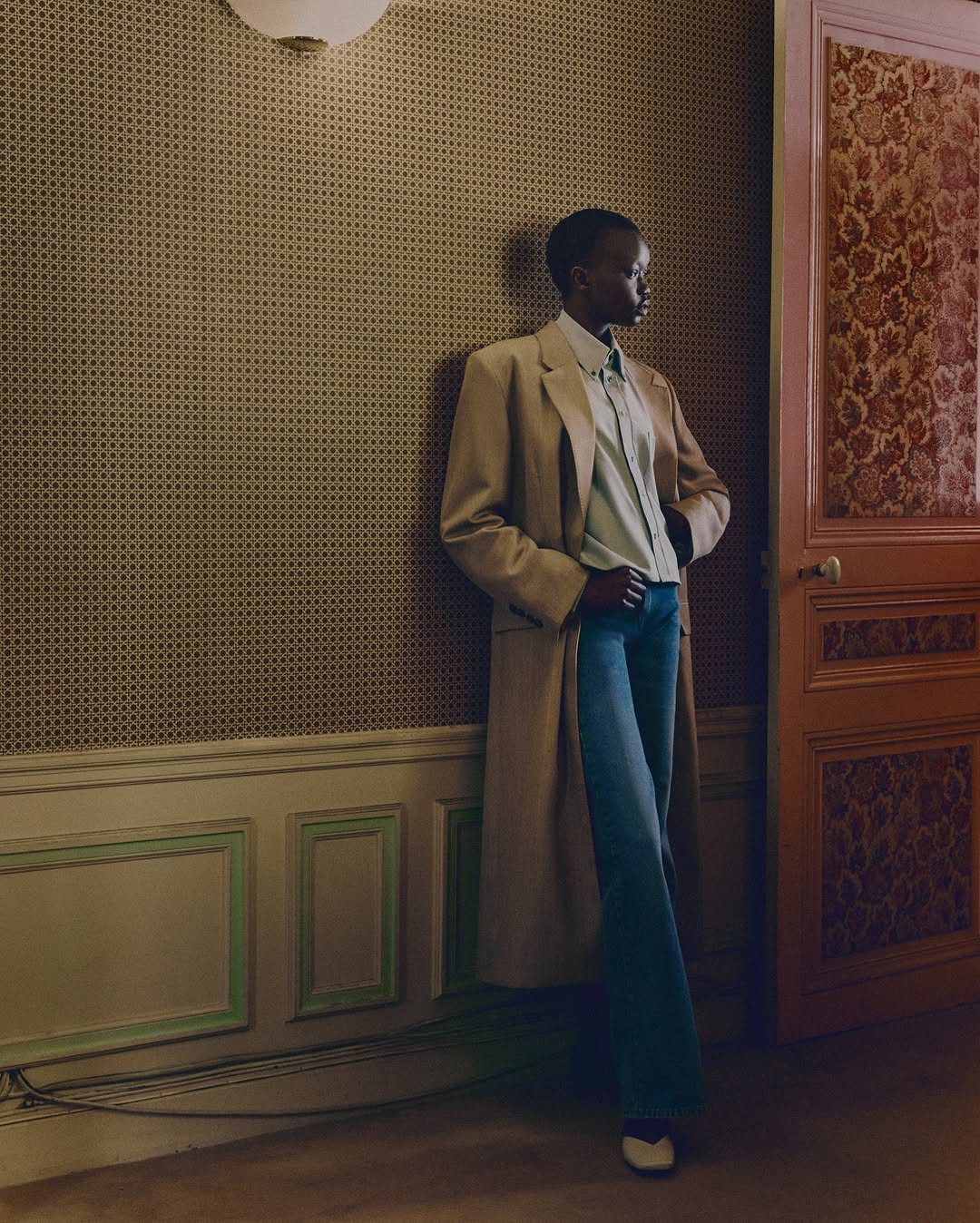 Model wearing a long beige coat and blue denim jeans standing against patterned wallpaper in a classic interior setting
