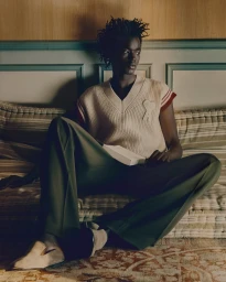 Young Black man seated indoors wearing a cream V-neck sweater vest and dark trousers against a paneled wall