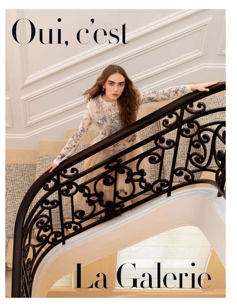 Woman in embellished gown poses on a staircase with black iron railing in a light-filled interior for a fashion or beauty editorial.