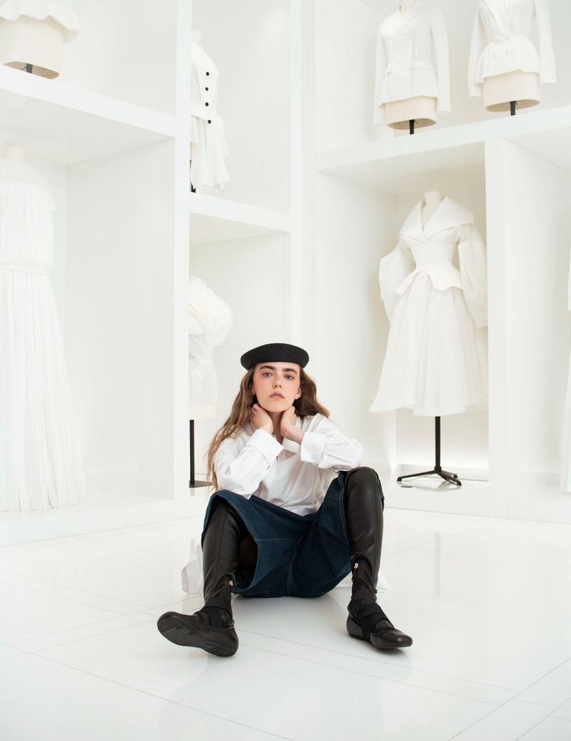 Woman in black beret and boots sits on a white floor with white dresses displayed on mannequins in a studio-like setting.