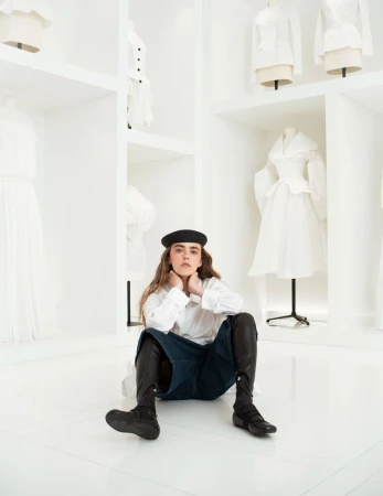 Woman in black beret and boots sits on a white floor with white dresses displayed on mannequins in a studio-like setting.