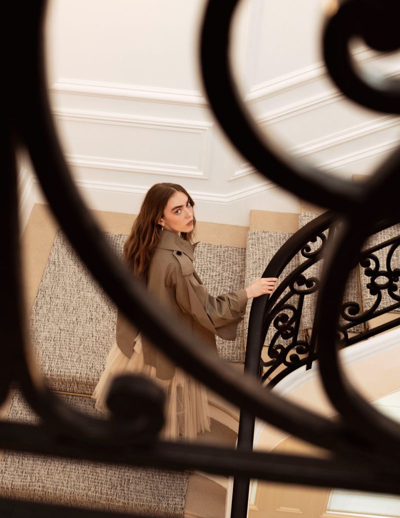 A woman walks up a staircase wearing a khaki trench coat and beige tutu skirt, holding the black wrought-iron banister.