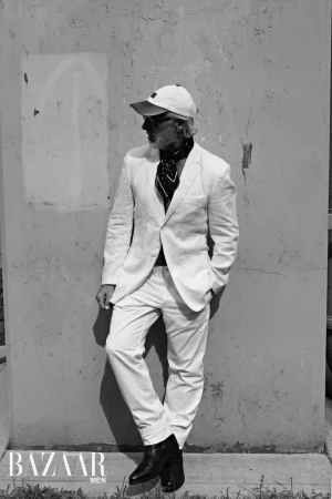 A stylish older man in a white suit leans against a weathered wall, showcasing a bandana and a white baseball cap in a black and white portrait.