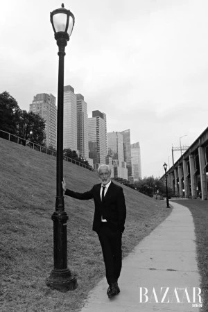 Black and white outdoor shot of an adult male in a suit, leaning on a vintage street lamp in a park with buildings in the background.