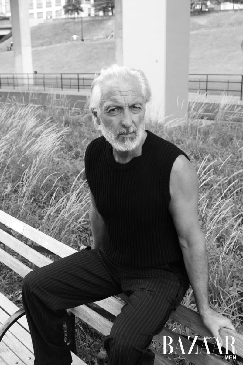 Black-and-white portrait of a mature adult male with gray hair, sitting on a park bench outdoors, wearing a dark sleeveless shirt and striped pants.