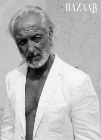 Monochrome portrait of an older adult male with gray hair, wearing a white blazer against a textured wall, fashion editorial style.