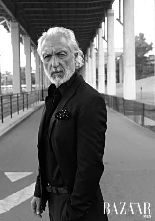 Black and white portrait of a mature man in a dark suit and button-down shirt under a bridge in an urban setting.