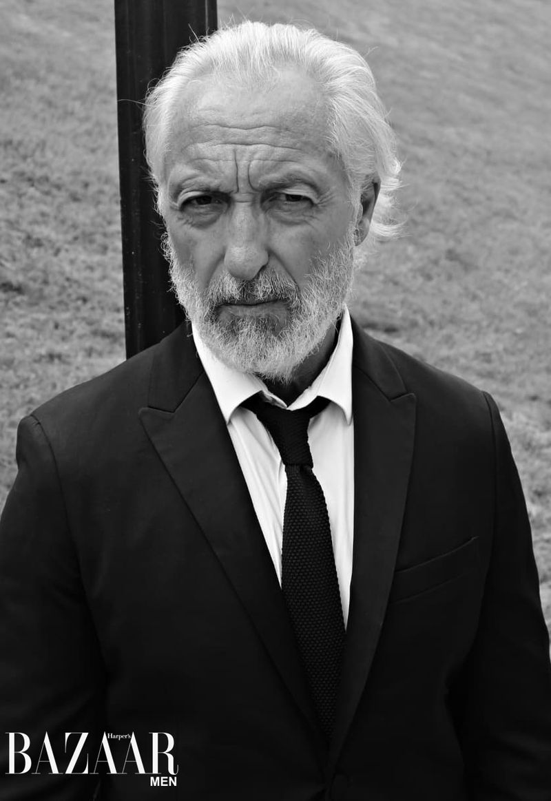 Black and white portrait of a mature man in a suit and tie, posing outdoors. He has gray hair, a beard, and a serious expression.
