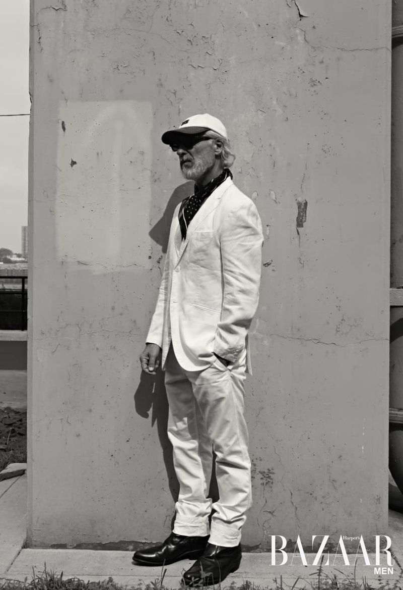 Older man in white suit posing against weathered wall for a fashion editorial. He's wearing a cap and neck scarf, with harsh shadows.