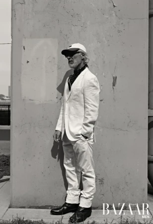 Older man in white suit posing against weathered wall for a fashion editorial. He's wearing a cap and neck scarf, with harsh shadows.