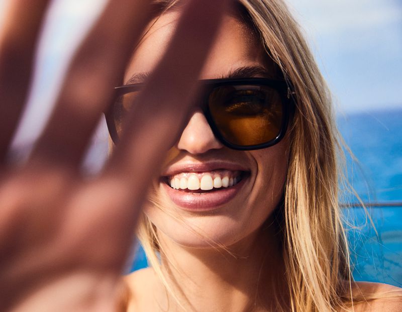 Smiling blonde woman in sunglasses holds hand up to camera. Close-up shot with blue water background, sunlit and bright mood.