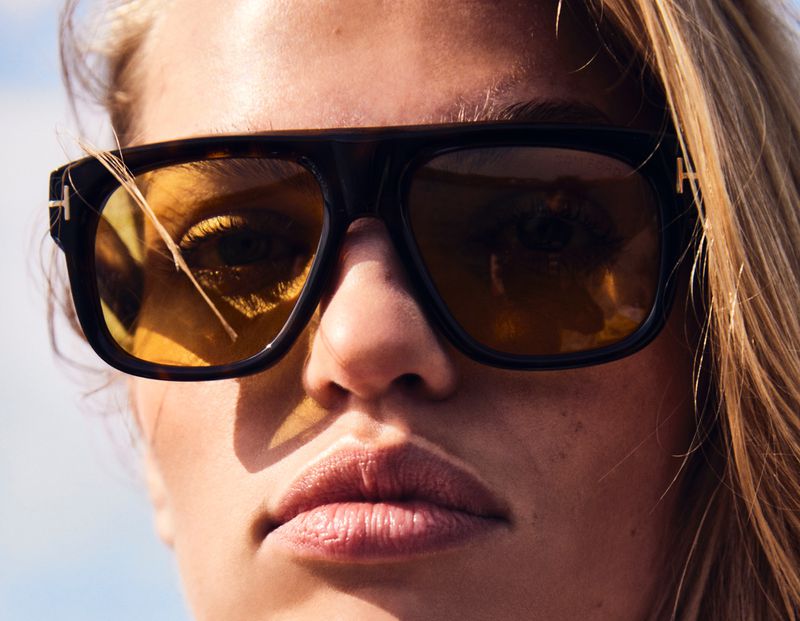 Close-up of a woman with light hair wearing brown and black sunglasses with yellow lenses reflecting the sky and the sun.