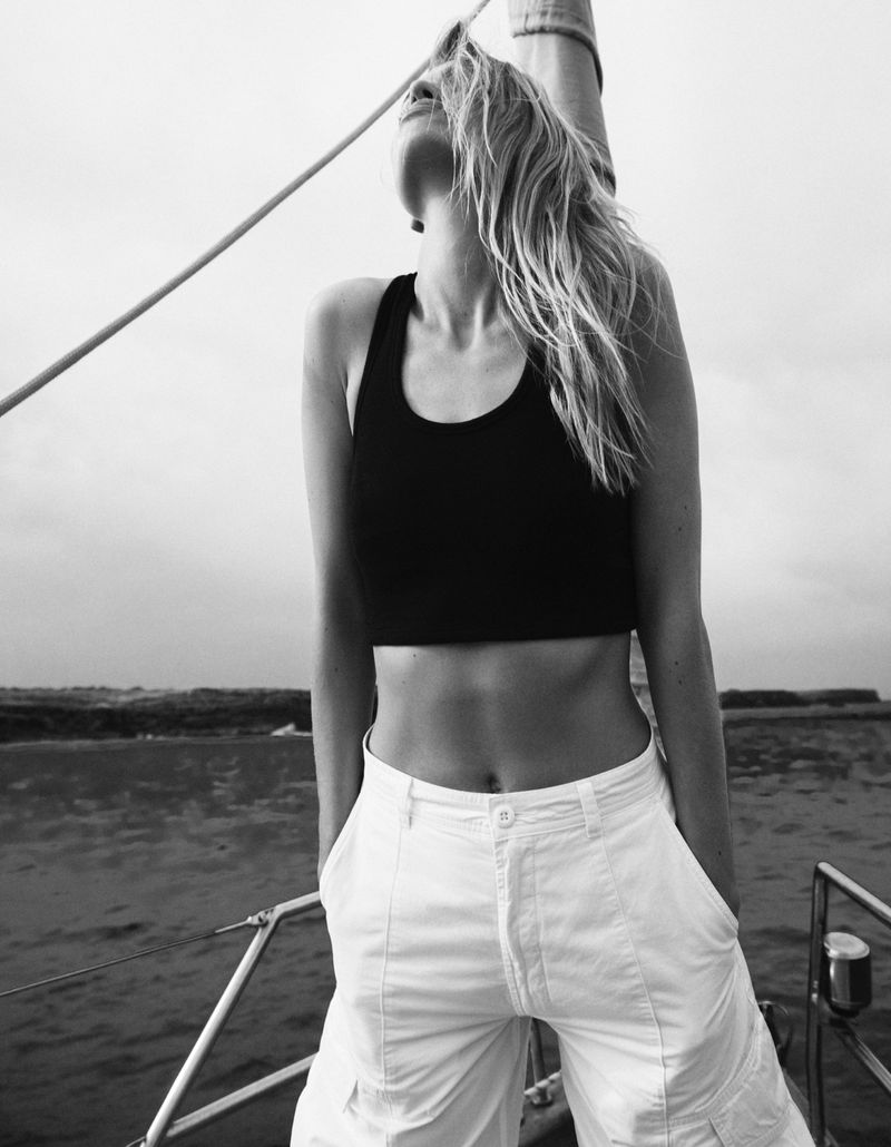 Woman in black crop top and white cargo pants stands on a sailboat looking up, with the ocean and sky in the background in grayscale.