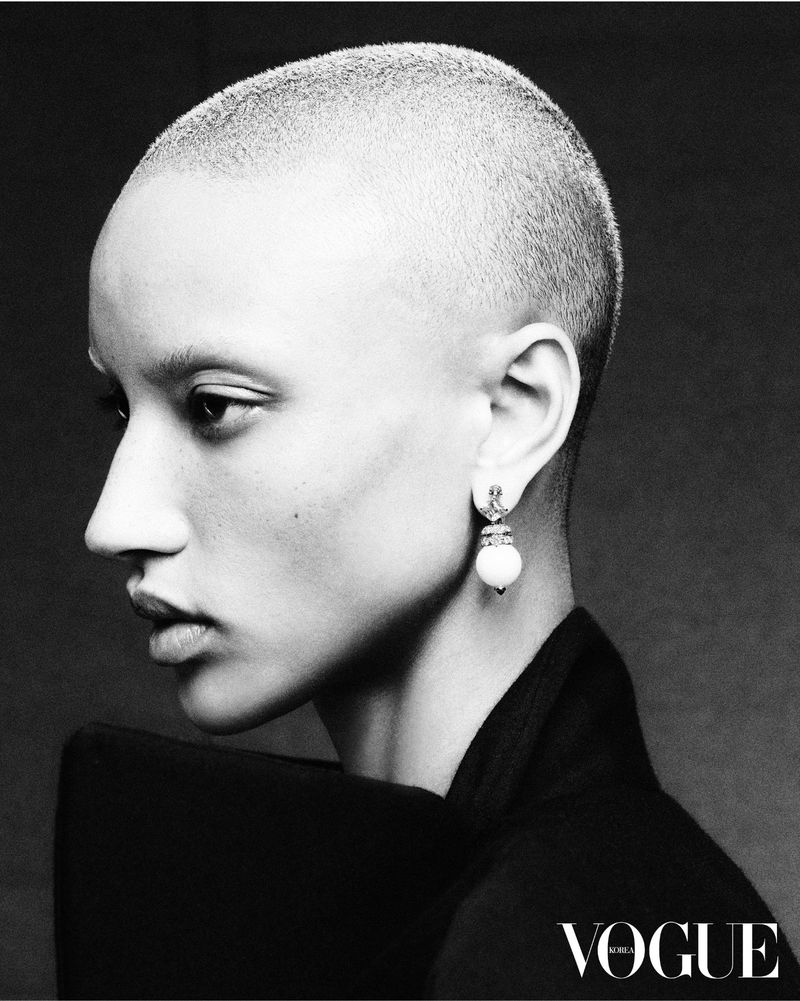 Black and white portrait of an adult female with a shaved head wearing jewelry and a black top, with the Vogue logo visible.