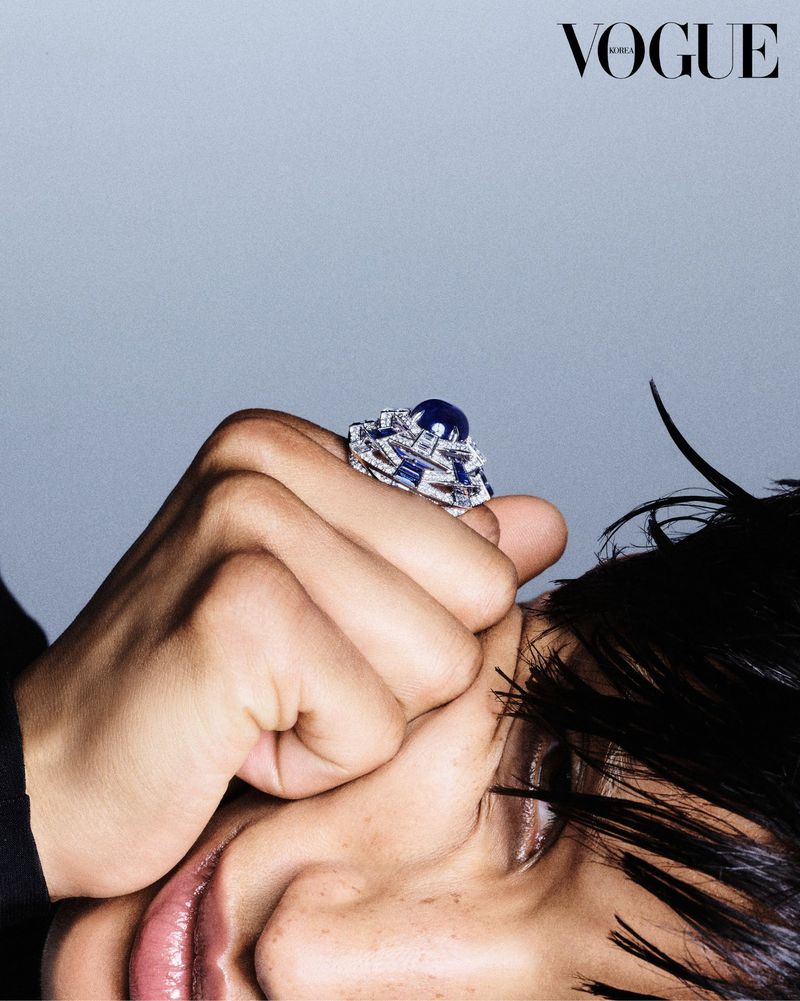 Close-up of a sapphire and diamond ring on a person's hand, with a neutral gray background and a 'Vogue' logo in the upper right corner.