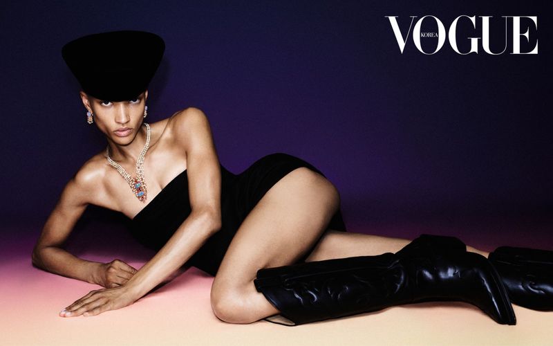Fashion shot featuring a woman lying down in a black strapless dress, hat, jewellery, and cowboy boots against a purple and orange gradient background.