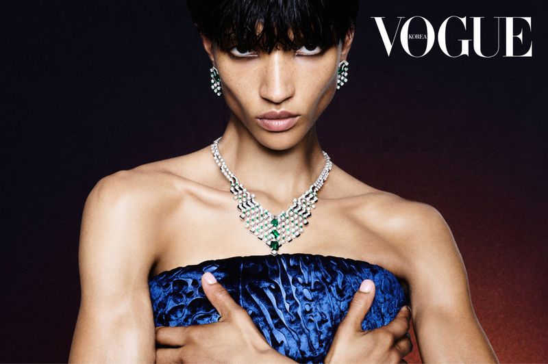 Close-up of a fashion model wearing diamond and emerald jewelry, a blue velvet dress, with VOGUE Korea logo visible in the upper corner.