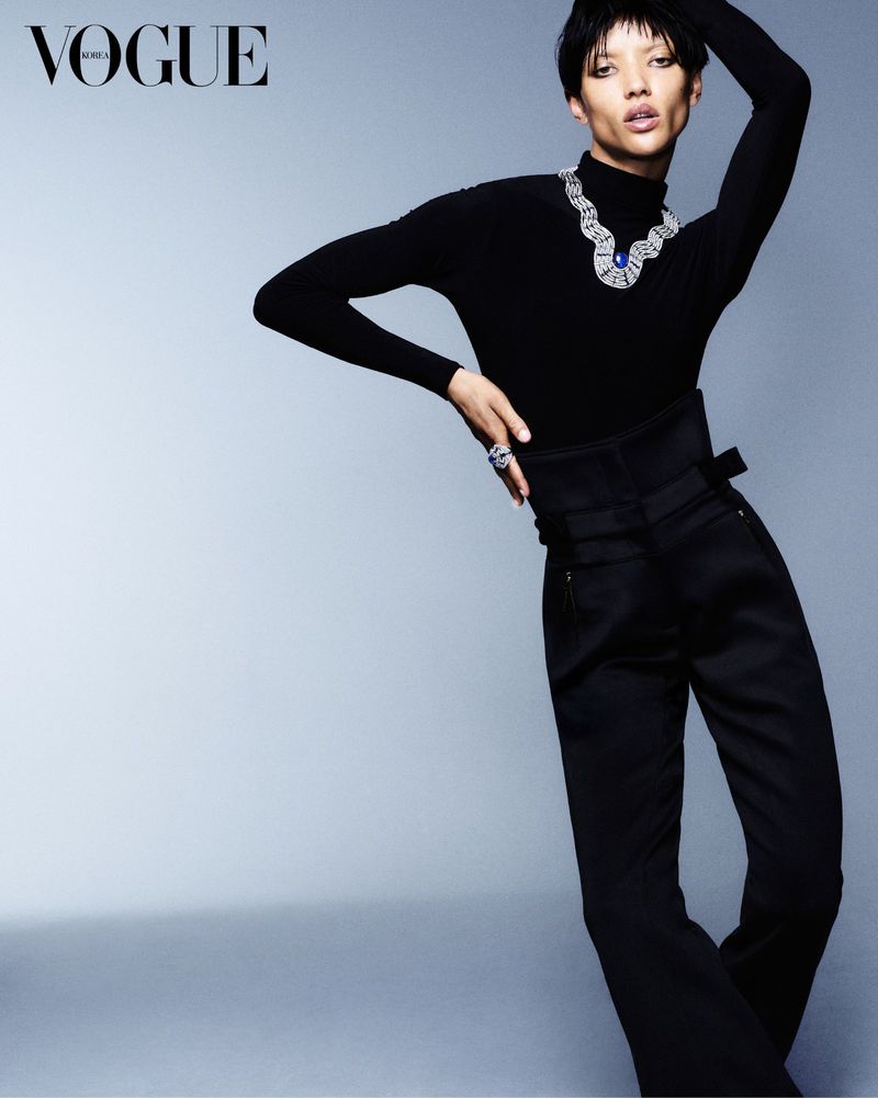 Fashion editorial shot of a stylish adult female model in a black turtleneck, high-waisted pants, and a large jeweled necklace posing in studio.