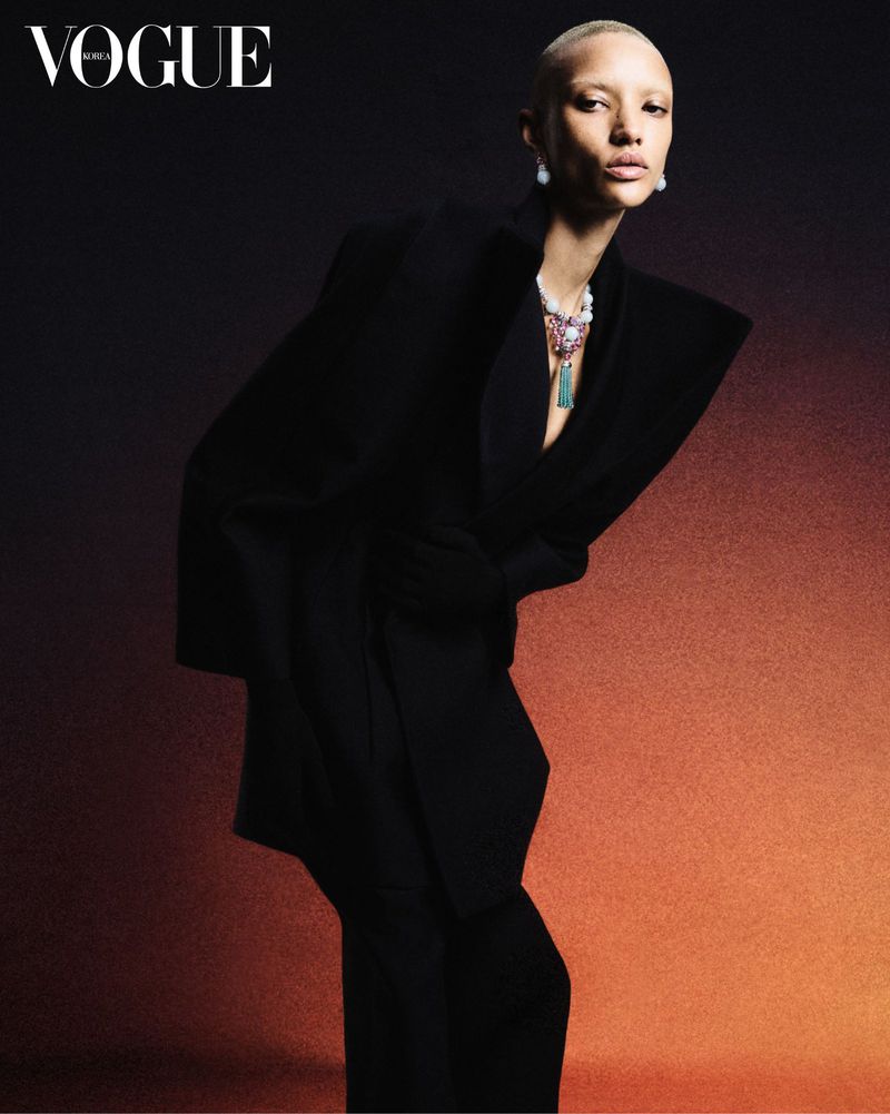 Fashion editorial shot of a bald woman in a black suit and ornate necklace, posing against a dark gradient background.