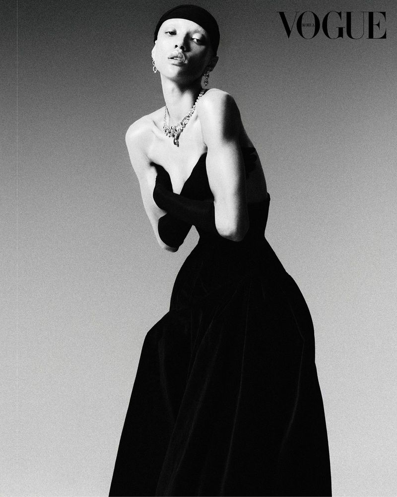 Black and white portrait of a model in a black dress with gloved hands, accessorized with a large necklace and earrings, Vogue logo.