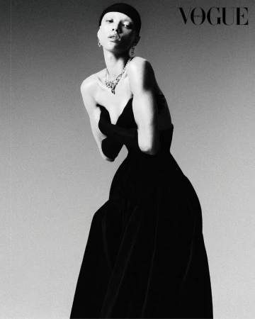 Black and white portrait of a model in a black dress with gloved hands, accessorized with a large necklace and earrings, Vogue logo.