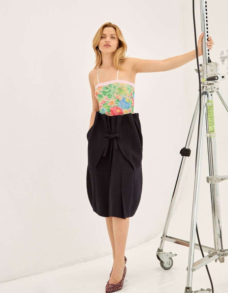 A blonde-haired woman poses in a floral top and black skirt in a photo studio, with a lighting rig visible in the background.