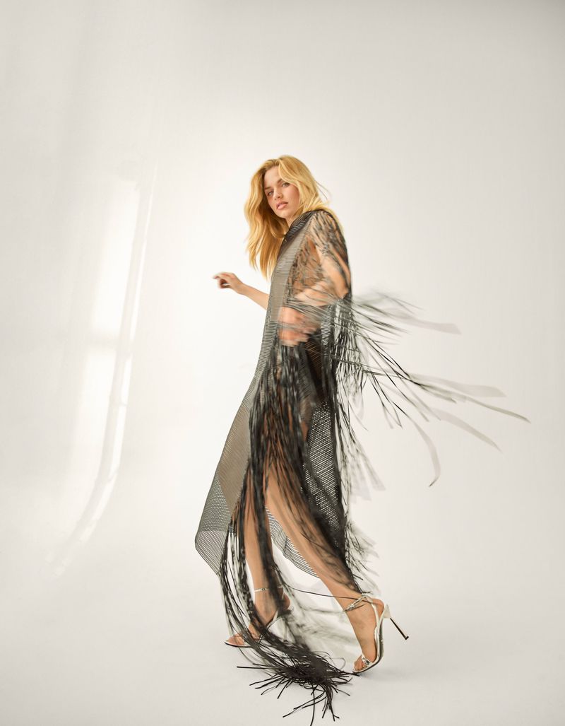 Fashionable woman in a fringed dress poses in a bright studio, showcasing a blend of elegance and avant-garde style with neutral tones.