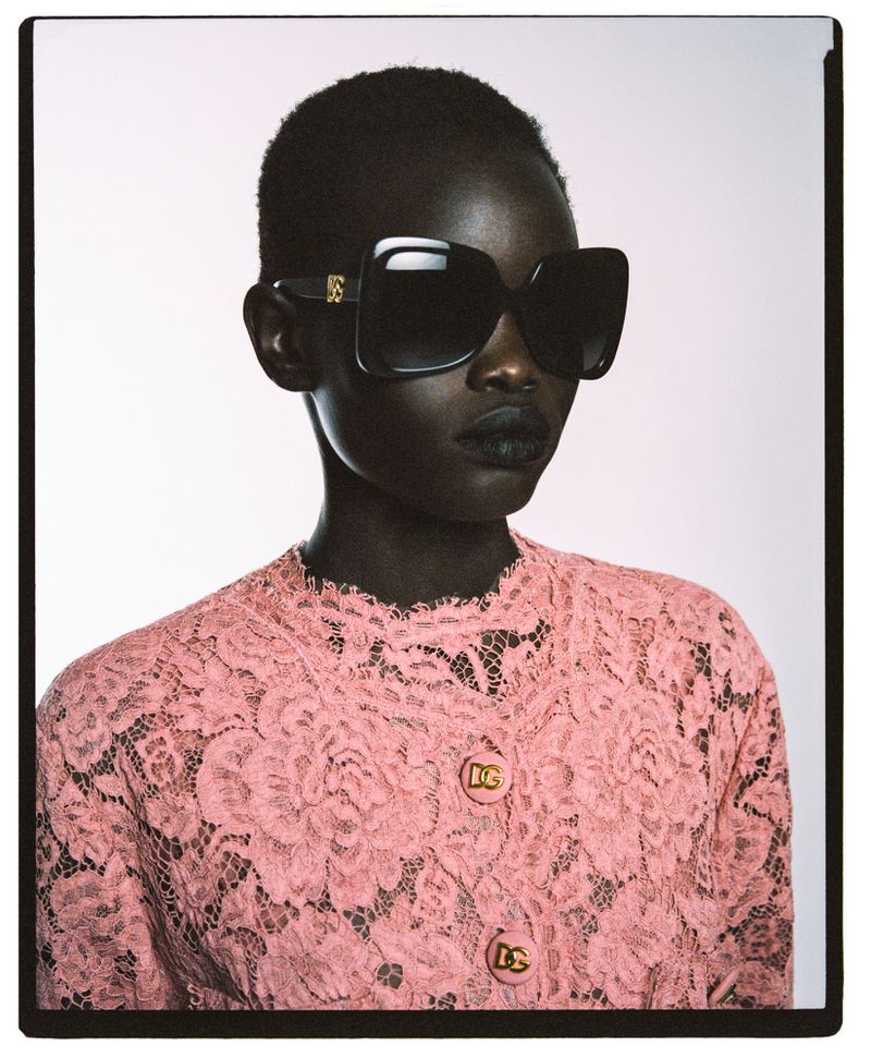 Portrait of an African woman wearing a pink lace jacket, oversized sunglasses, and dark lipstick against a light background.