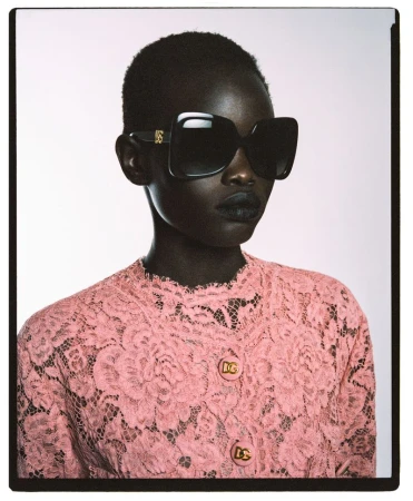 Portrait of an African woman wearing a pink lace jacket, oversized sunglasses, and dark lipstick against a light background.