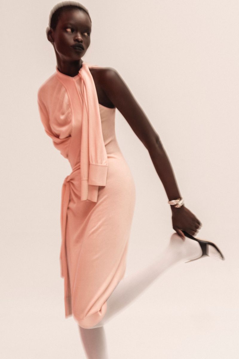 Fashionable dark-skinned woman poses in a stylish peach dress, holding a high heel shoe against a plain backdrop in a studio setting.