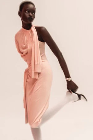 Fashionable dark-skinned woman poses in a stylish peach dress, holding a high heel shoe against a plain backdrop in a studio setting.