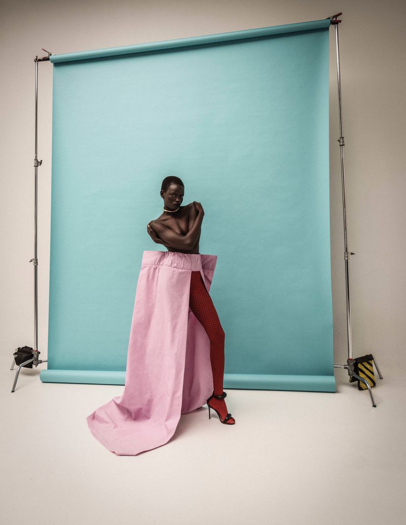 A Black fashion model in a modern skirt stands in front of a studio backdrop, posing in red fishnet stockings and black high heels.