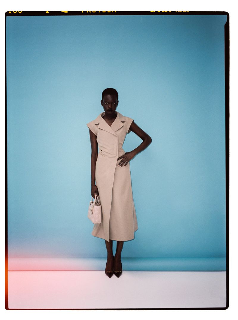 Elegant Black woman in beige trench dress and black heels holds purse against a gradient blue backdrop in a fashion studio shot.