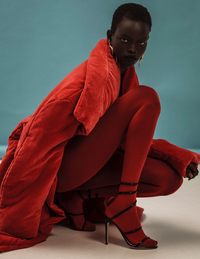 Fashionable woman in a squatting pose wearing a red coat, red tights, and high-heeled strappy sandals against a blue background.