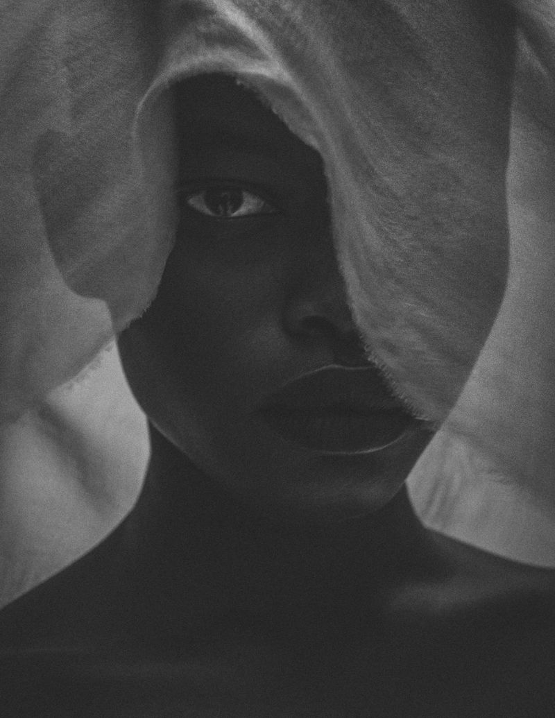 Black and white portrait of a Black woman with fabric draped over her head, partially covering her face in a studio setting.