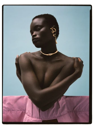 Close-up studio shot of a woman with dark skin wearing gold jewellery, arms crossed, against a light blue backdrop and pink fabric.