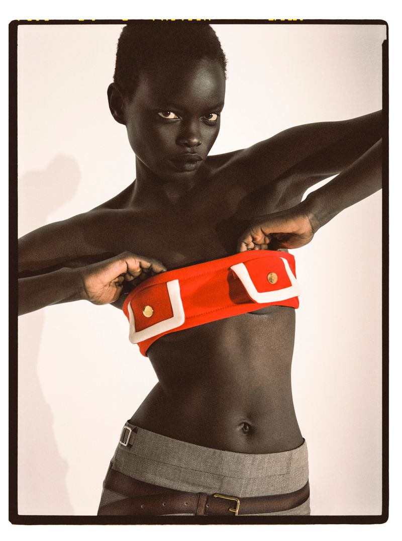 Adult female fashion model poses indoors wearing a red tube top with white trim and grey pants with brown belt against a white backdrop.