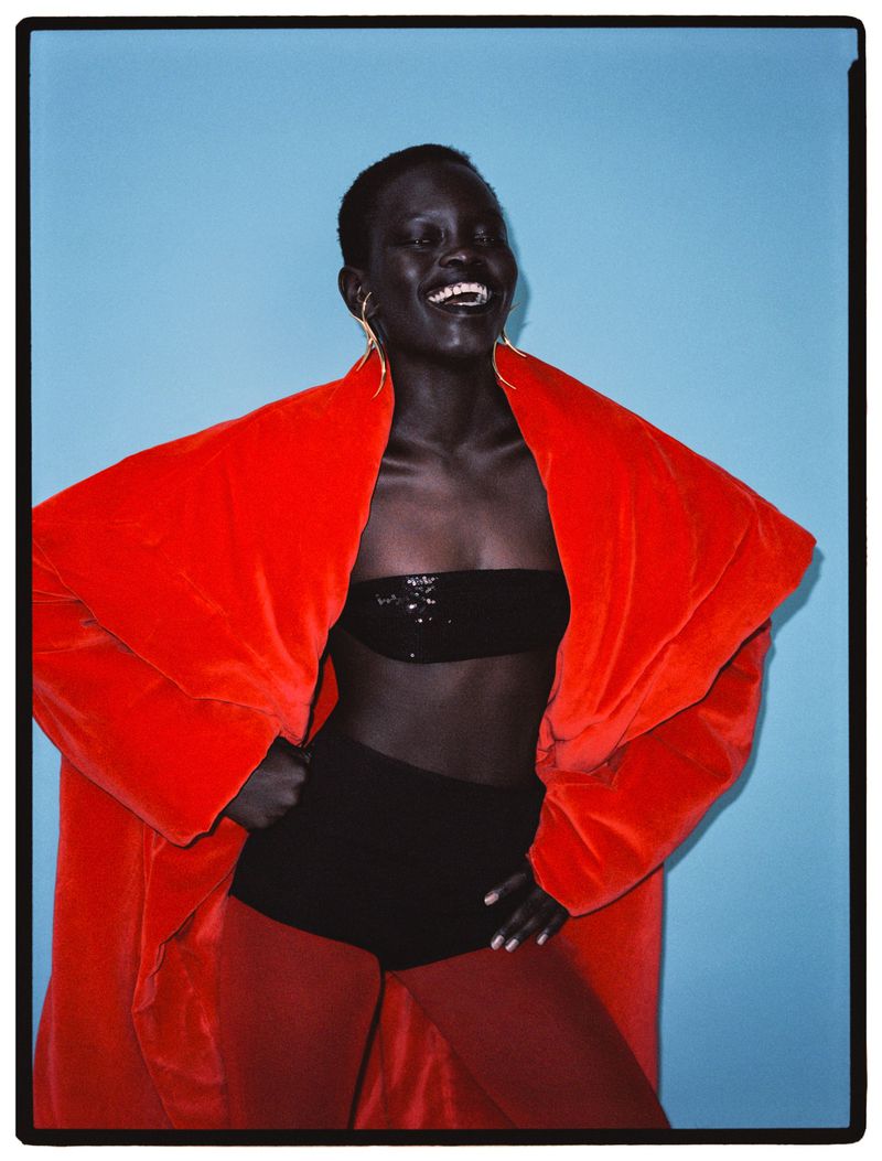 Fashionable portrait of a smiling dark-skinned woman in an orange coat, black top and shorts, posing against a bright blue backdrop.