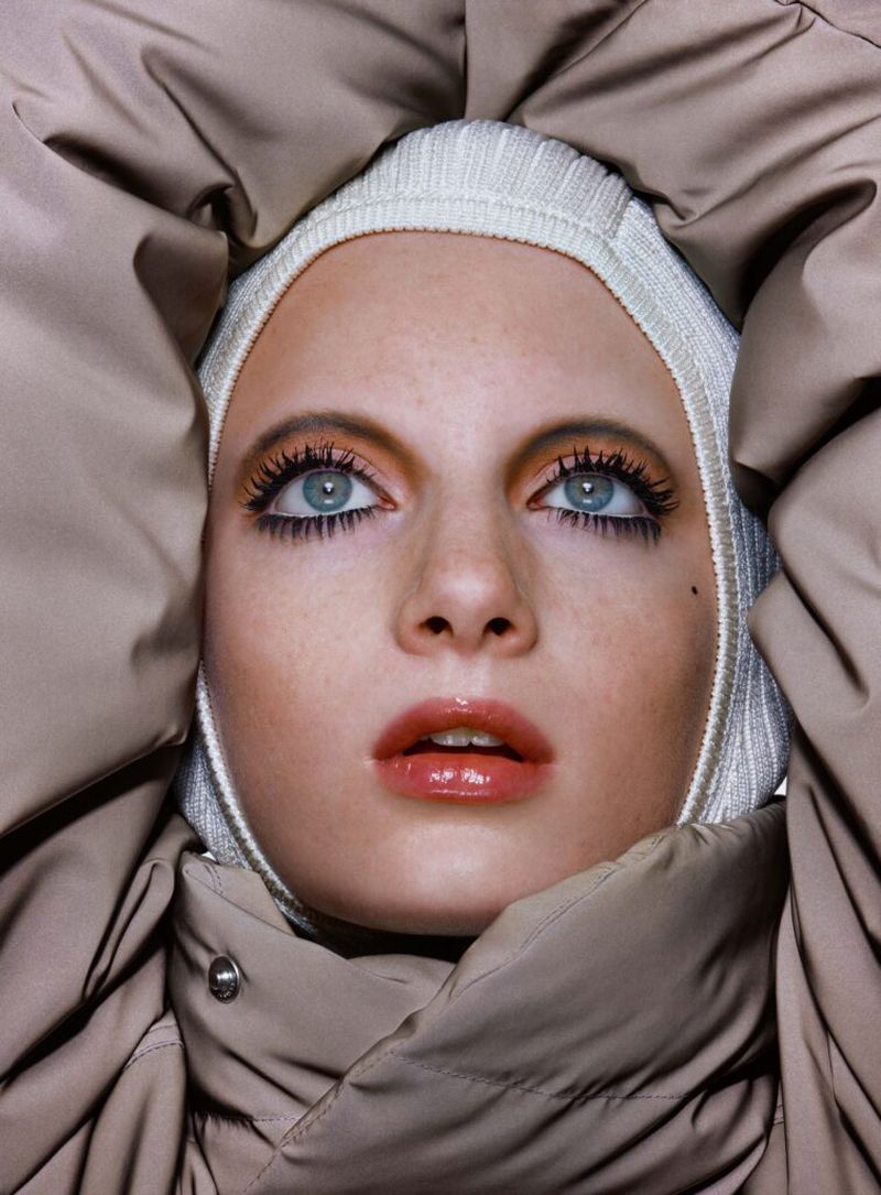 Close-up portrait of a fair-skinned young woman with blue eyes, wearing a knit hat and a beige puffer jacket around her face.
