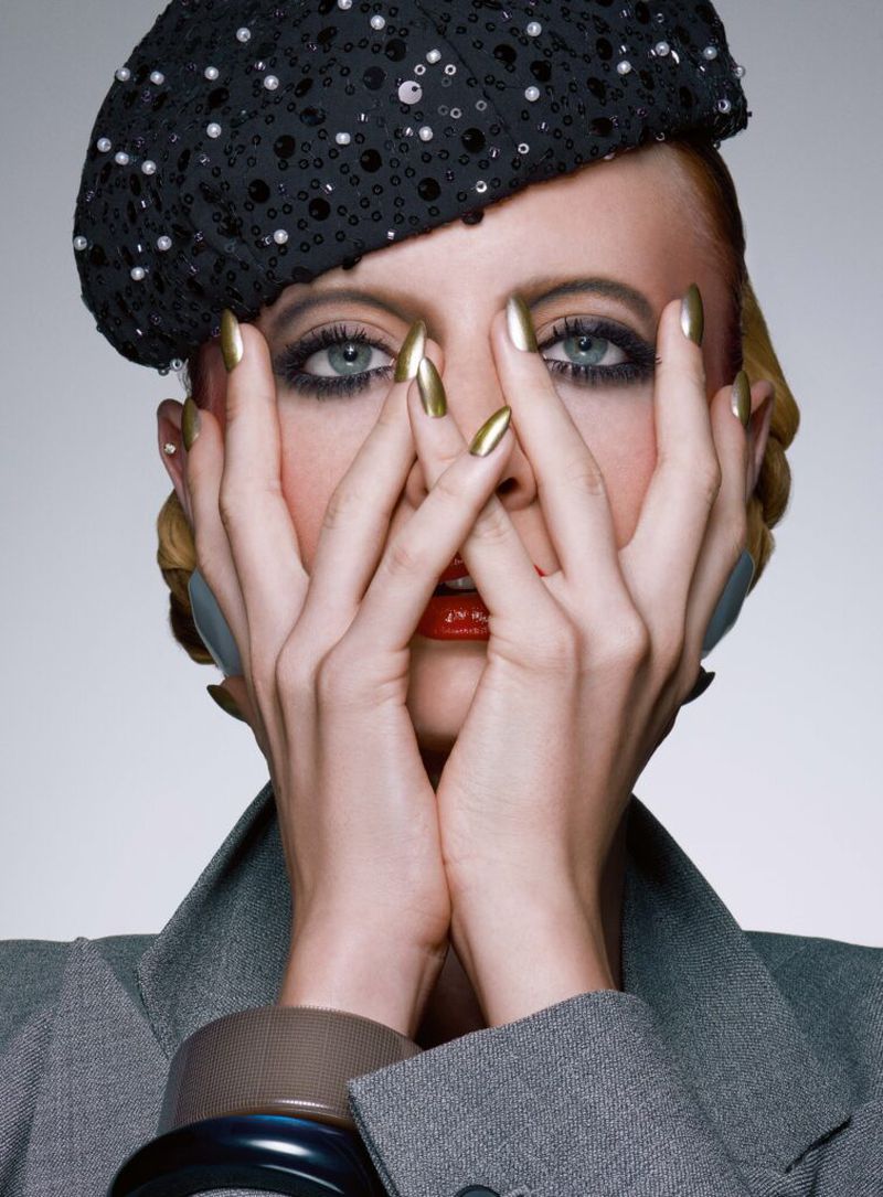 Close-up portrait of a stylish adult female model with blue eyes wearing a jeweled hat, grey jacket, and golden fingernails holding her hands up.