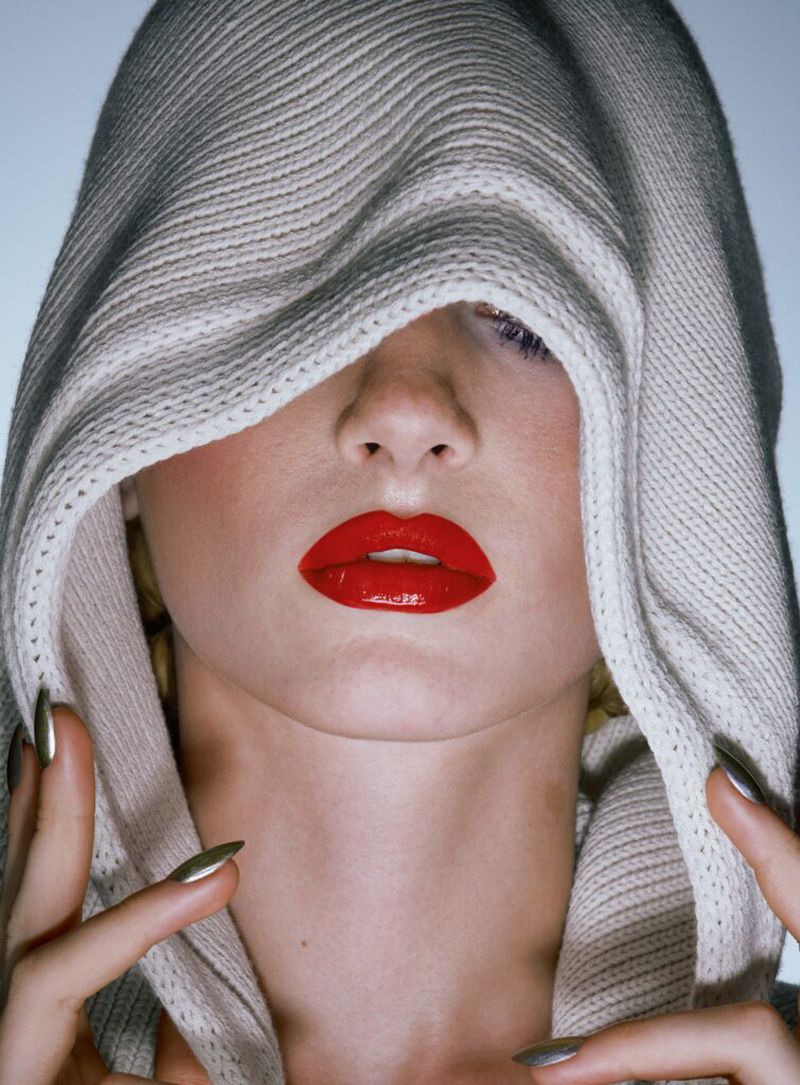 Close-up of a model wearing a knitted hood, bright red lipstick, and long silver nails in a studio setting with a pale blue background.