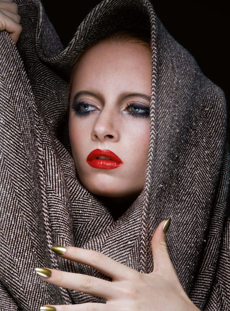 Woman in herringbone fabric hood with red lips and gold nails poses against dark backdrop in beauty shot, eye contact to the left.
