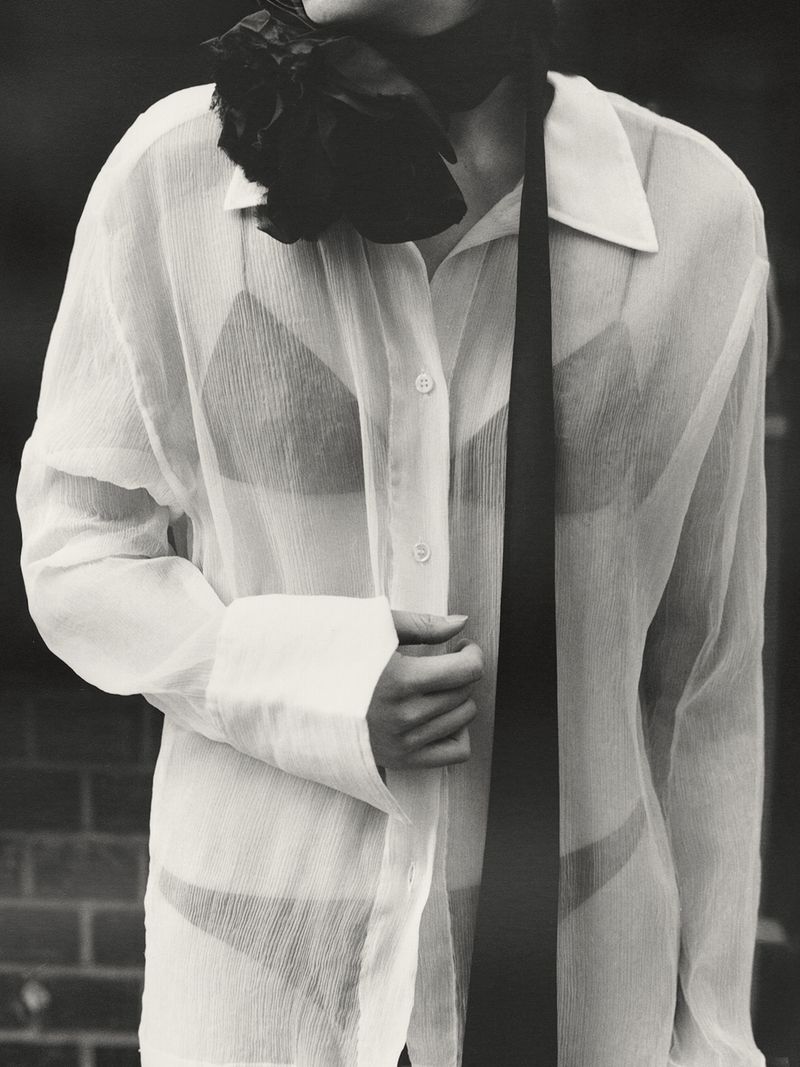 Monochromatic portrait of a woman in an unbuttoned, semi-sheer button-down shirt with a black bra and tie, set against a brick wall.