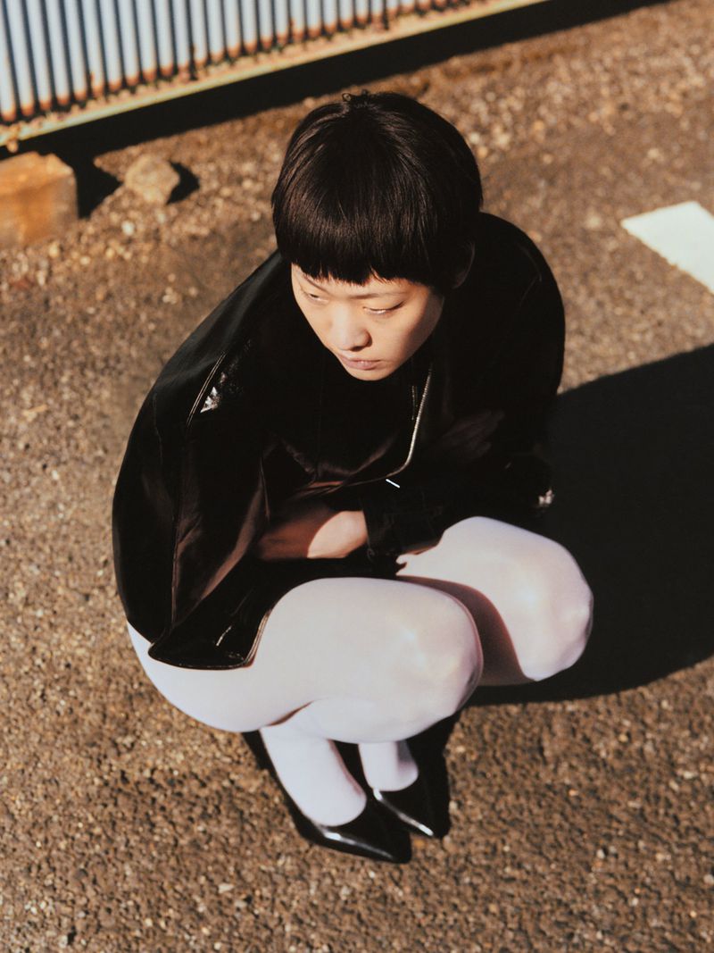 Asian woman crouching on pavement wearing black top, white tights, and patent-leather shoes; natural light with slight shadows.