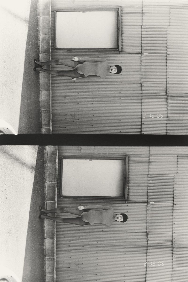 Monochromatic portrait of a woman in a swimsuit sitting against a corrugated metal wall with framed windows, creating a vintage aesthetic.